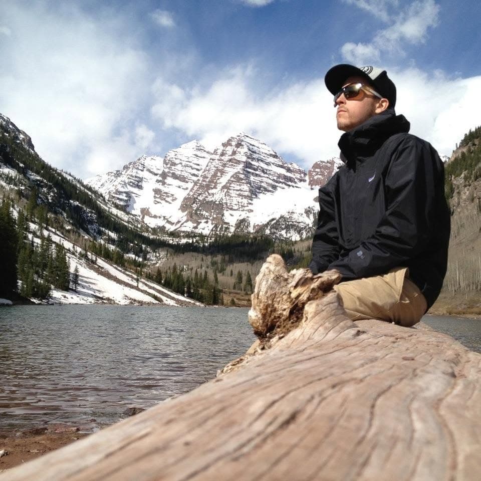 Brent P. Evans sitting on a fallen log beside an alpine lake, below snow-covered peaks